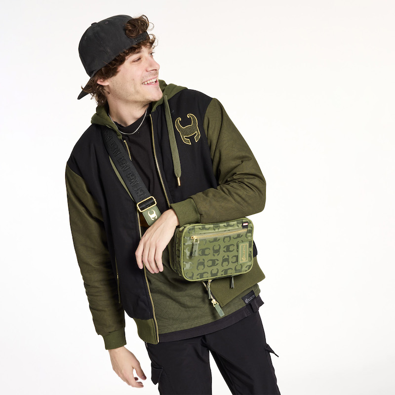 Man wearing the Loungefly COLLECTIV Marvel Loki The WEEKENDR Hooded Jacket and a backward black cap, wearing the Loungefly COLLECTIV The INFLUENCR Convertible Crossbody Bag, featuring an all over print of Loki's helmet 